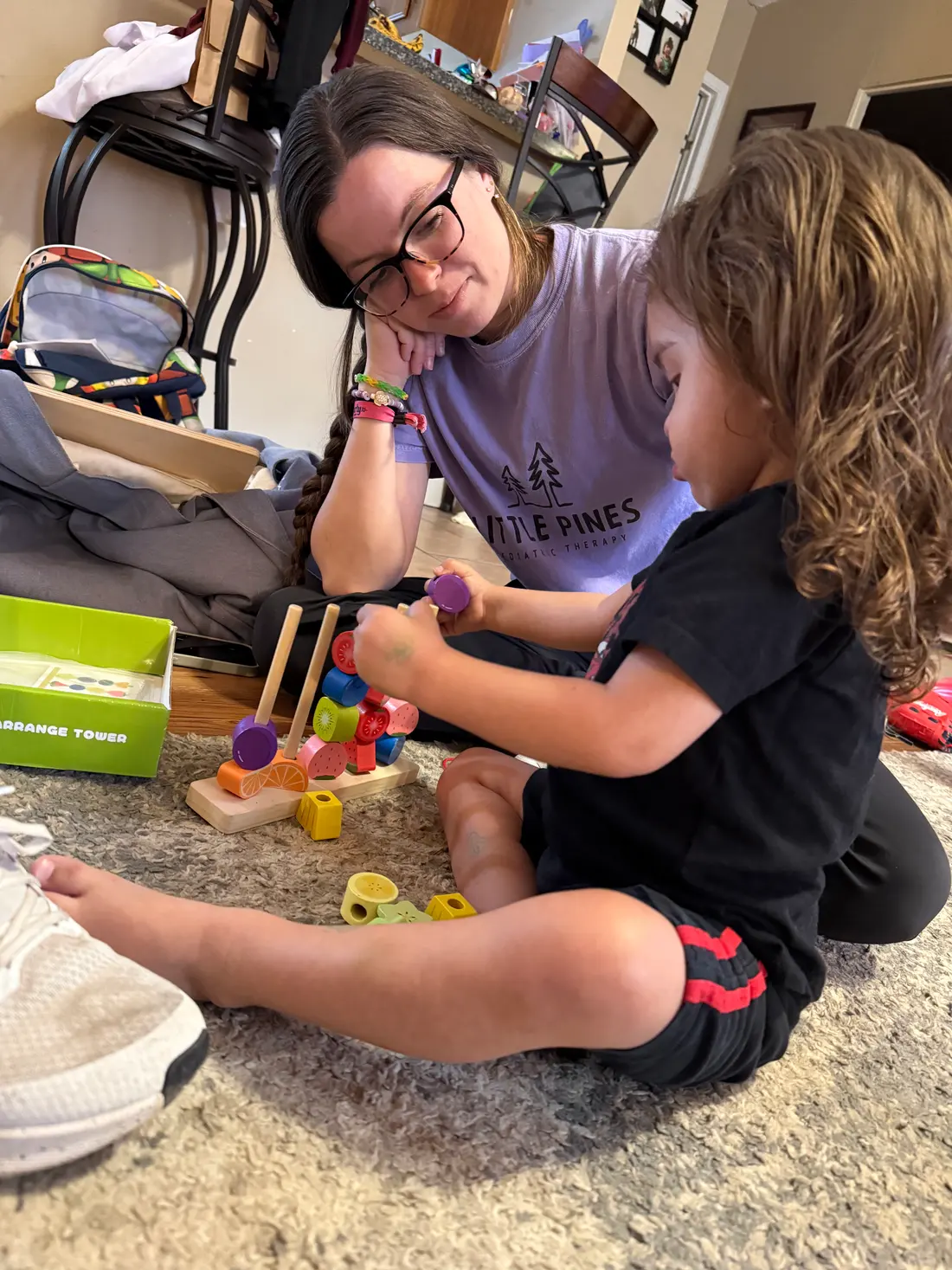 A Little Pines therapist sits on the floor beside a toddler, both stacking colorful wooden rings on a peg tower.