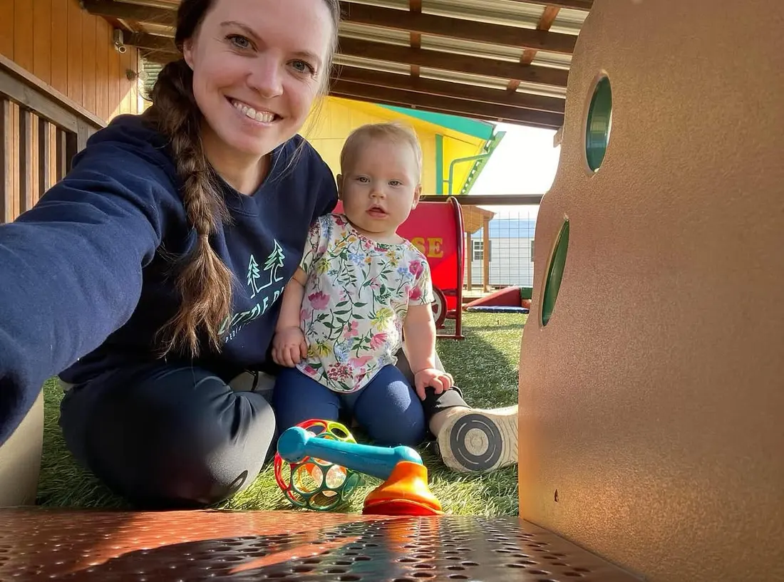 A therapist in a Little Pines sweatshirt sits under a playground structure with a baby who is reaching for a rattle.