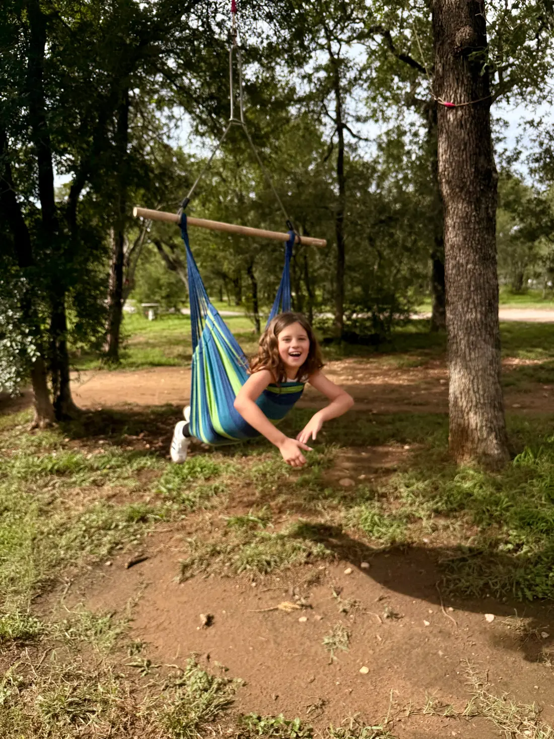 A girl leans forward laughing from a striped hammock swing hung between oak trees.