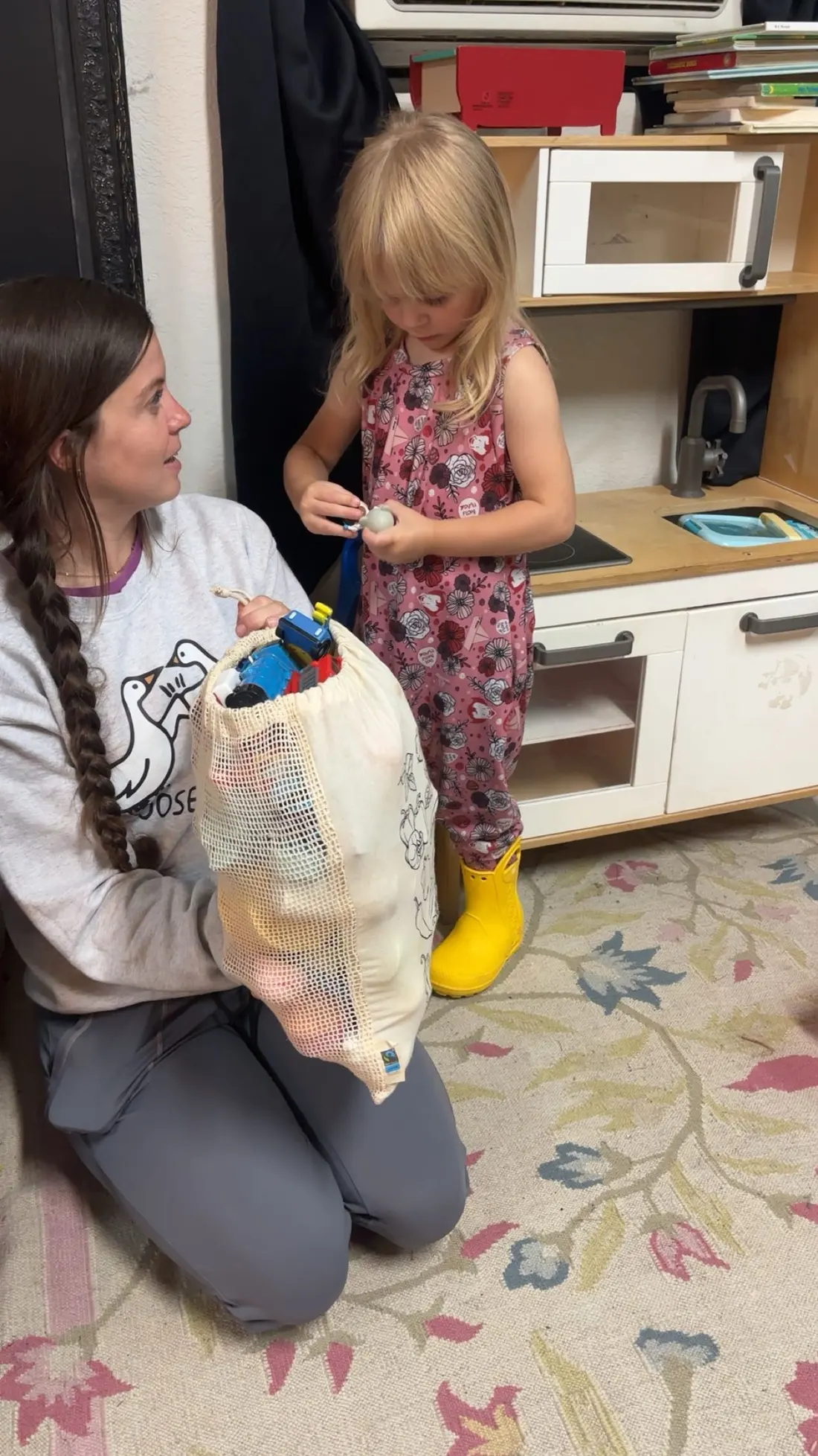 A therapist kneels with a mesh bag of toys while a toddler in yellow rain boots plays at a toy kitchen.