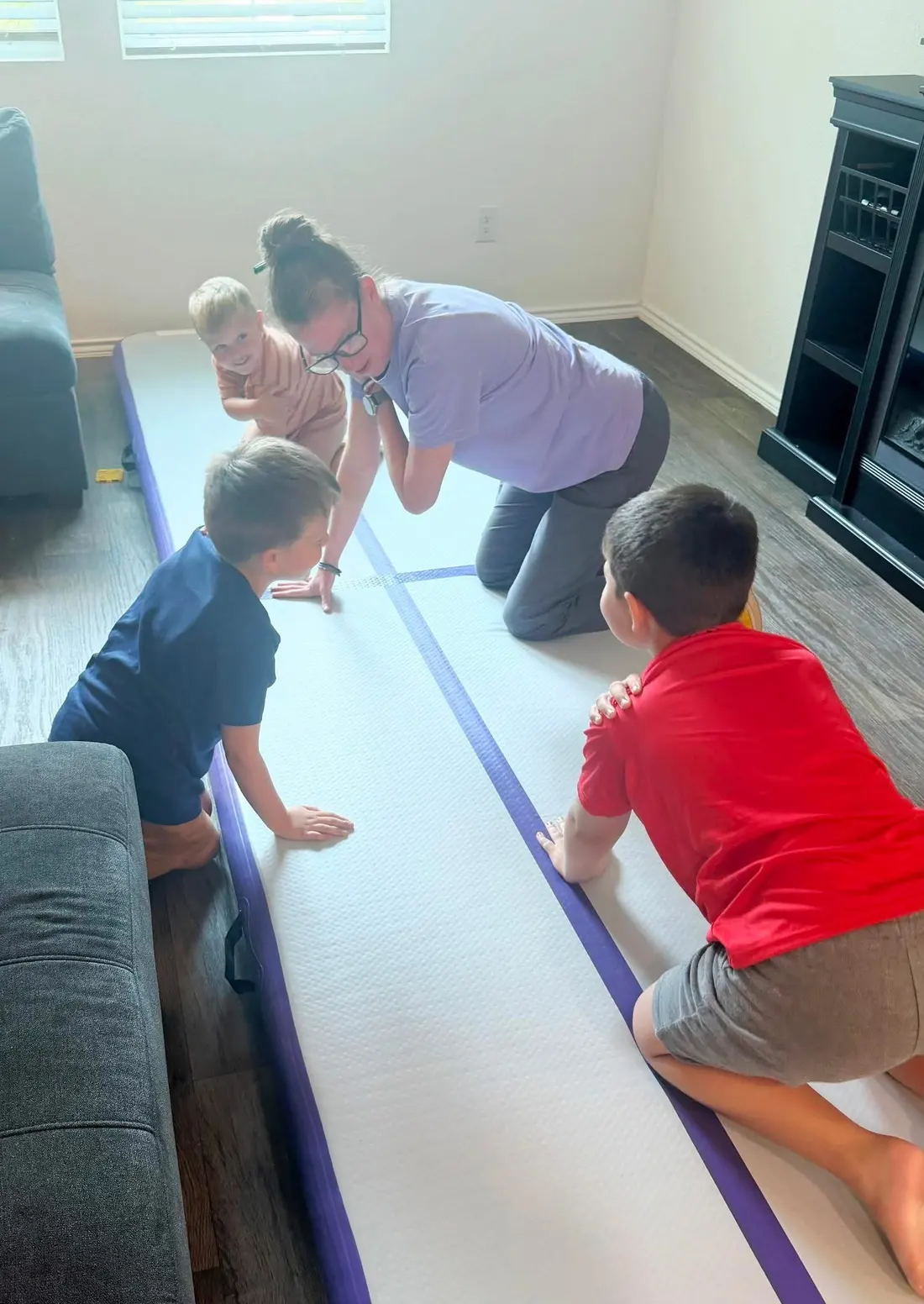 A therapist crouches at the end of a long padded floor mat as three children crawl toward her.