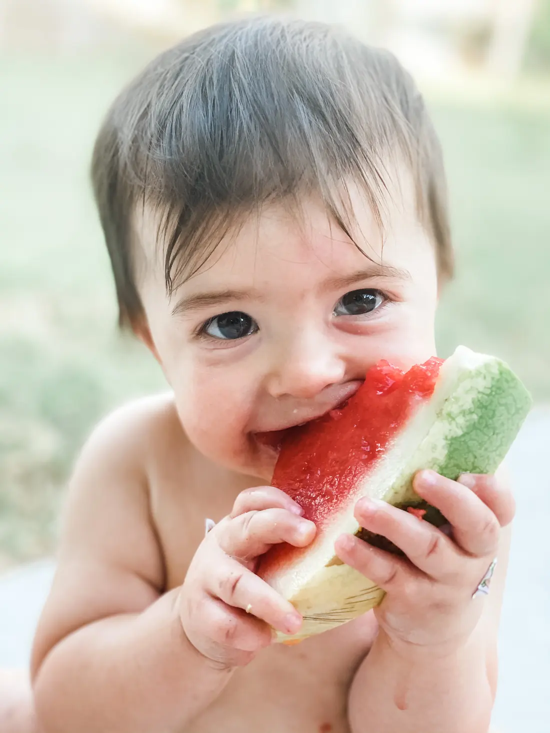 A baby holds a slice of watermelon with both hands and takes a bite.