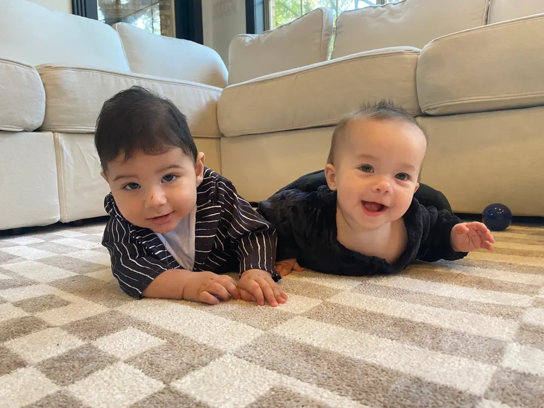 Two babies lie on their tummies on a living room rug, both propped on their forearms and smiling.