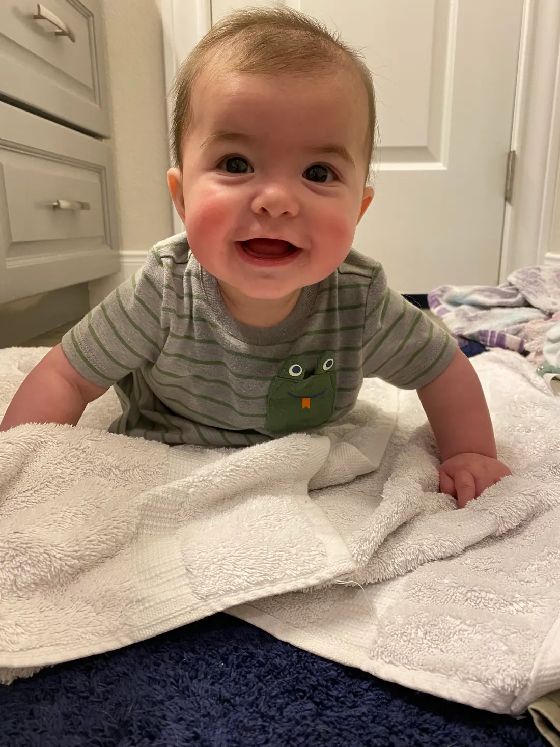 A baby pushes up onto their hands during tummy time on a folded towel, beaming at the camera.