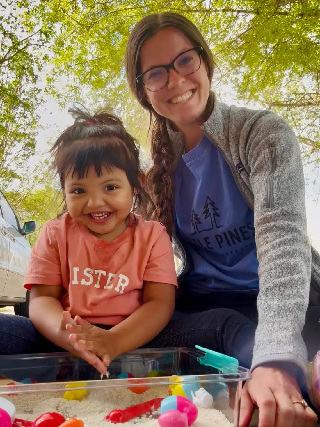 Shannon, in a Little Pines Pediatric Therapy shirt, sitting outdoors beside a smiling toddler at a rice sensory bin.
