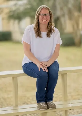 Stephanie smiling, seated on a wooden fence rail outdoors on a bright day.